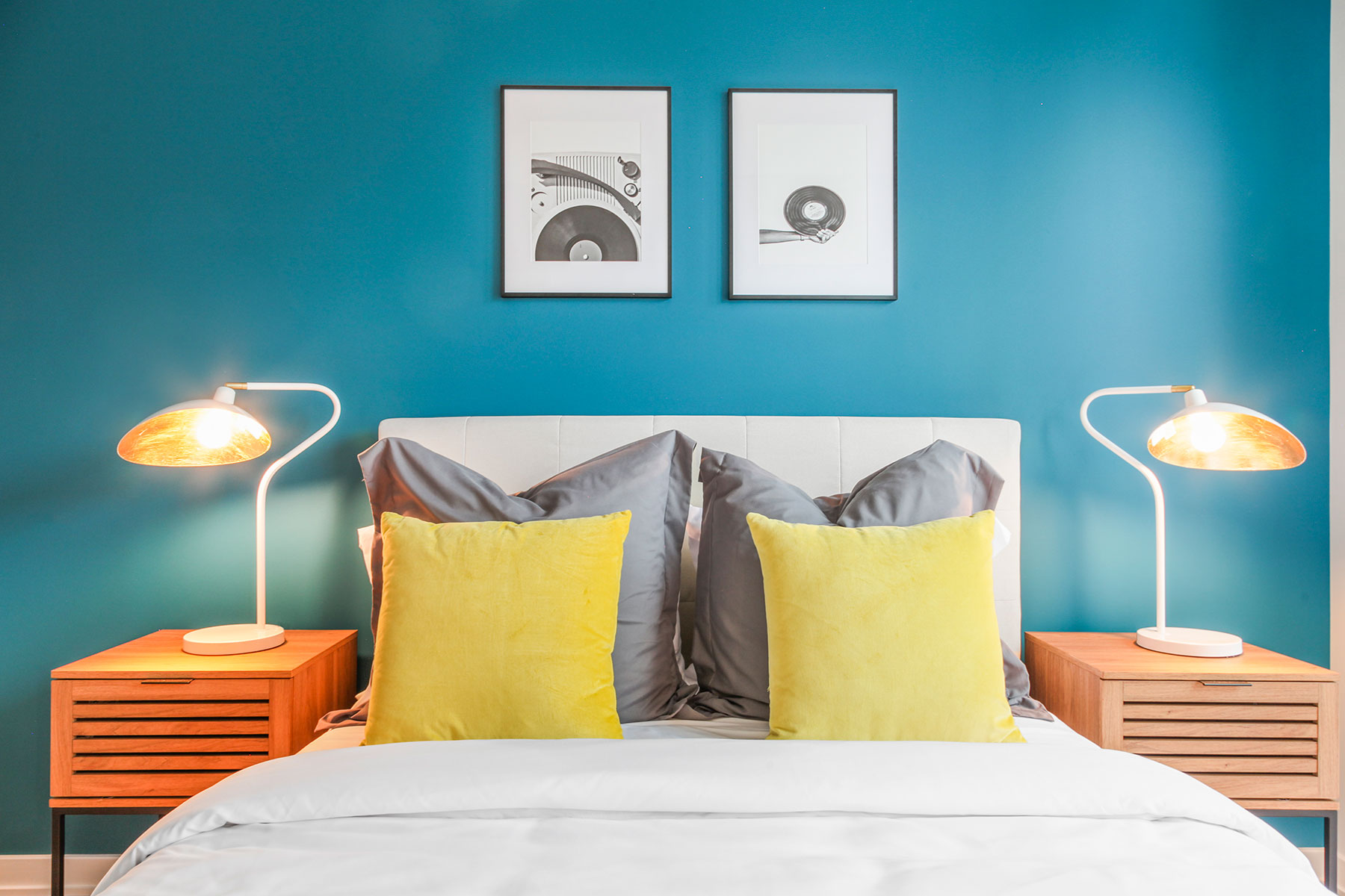 Detail of bed with modern bedding, accent pillows, fabric headboard, teal accent wall with framed art, matching wood and metal nighstands with lamps on each side
