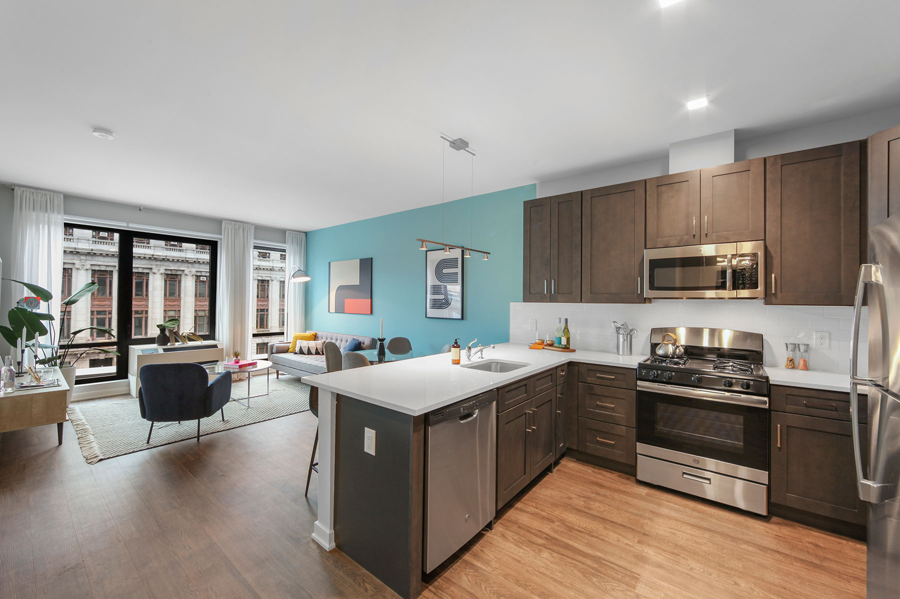 Bright modern kitchen with light counters, dark wood cabinetry, stainless steel appliances and modern fixtures and wood floors opening up to bright living room with large windows